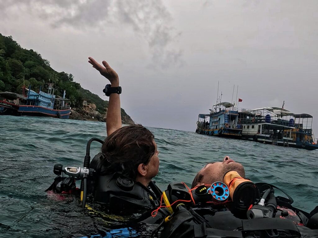 Two scuba divers during rescue training in Koh Tao. One diver tows an unresponsive diver at the surface while raising an arm to signal for help, with dive boats and coastline in the background.